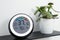 Digital hygrometer with thermometer and green plant on shelf, closeup