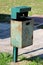 Dilapidated partially rusted green metal trash can with faded color mounted on metal pole with concrete foundation next to stone