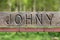 Dilapidated vintage retro sign with carved Johny on top mounted on metal pole of wire fence with green garden in background