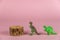 The dinosaur miniatures stand next to the pine pedestal against the pink background. Small green figures of predatory dinosaurs