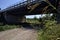 Dirt path next to a brook bordered by trees and reeds with a viaduct passing over it in the italian countryside