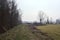 Dirt path next to a forest by the edge of a field with a bell tower in the distance framed by a tree on a cloudy day