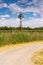 A dirt road through a green field / farm with a windmill and cloudy sky