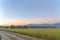 Dirt road and vast grassy field with houses and mountain background at sunset