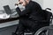 Disabled businessman sitting at workplace in wheelchair and holding hand on forehead