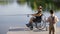 Disabled fisherman on the wheelchair fishing on the local lake on the pier and the the bucket is on the pier and his son