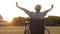 Disabled woman sitting in wheelchair on field against background sunset, raises his hands up and spreads them on sides