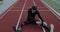 Disabled young man with prosthetic running blades stretching back while sitting at track on sports field. Male person