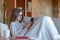 Disapointed young pretty brunette woman looking at the smartphone sitting on the sofa at home covered with a blanket. She wears gl