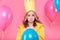 Disappointed young woman in party hat surrounded by colorful balloons, isolated over pastel pink background.