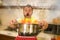 Disaster home cook at kitchen- young funny and desperate man in cooking apron holding pot in flames in stress and fear making a