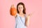 A disgruntled adult woman holds a pumpkin at arm`s length standing on a pink background. Selective focus