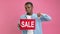 Disgruntled black teenager in denim shirt looks at sale banner and points his finger down on pink studio background. Bad