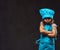 Disgruntled little girl dressed in blue cook uniform standing with crossed arms. on dark textured background.