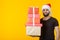 Disgruntled young man with a beard in a Santa Claus hat holds five gift boxes posing on a yellow background with