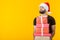 Disgruntled young man with a beard in a Santa Claus hat holds five gift boxes posing on a yellow background with