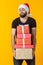 Disgruntled young man with a beard in a Santa Claus hat holds five gift boxes posing on a yellow background. Concept of