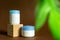 Display wooden podium with crem jars on a brown background with green leaves and shadows. Face care containers. Cosmetics mockup.