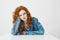 Displeased disappointed young girl with red curly hair sitting at table over white background