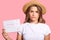 Displeased European woman in straw hat and casual t shirt, holds menstruation calendar, wears casual t shirt, isolated over pink