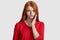 Displeased red haired woman with freckled skin, keeps hand on cheek, suffers from toothache, has sensitivity, wears casual red