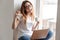 Displeased woman in t-shirt and eyeglasses sitting on windowsill