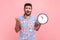 Displeased young adult nervous bearded man with dark hair screaming, showing wall clock at display, deadline, wearing blue casual