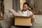 Dissatisfied black male customer opening box from online store, taking out packaging, unhappy with received item at home