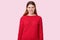 Dissatisfied dark haired woman clenches teeth, frowns face with annoyance, dressed in red sweater, models over rosy background.