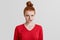 Dissatisfied pleasant looking ginger woman with hair knot curves lips in dissatisfaction, has grumpy expression after quarrel with