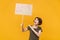 Dissatisfied young protesting woman pointing hand on protest sign broadsheet placard on stick isolated on yellow