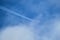 Distant passenger jet plane flying on high altitude through white clouds on blue sky leaving white smoke trace of