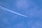 Distant passenger jet plane flying on high altitude through white clouds on blue sky leaving white smoke trace of