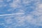 Distant passenger jet plane flying on high altitude through white clouds on blue sky leaving white smoke trace of