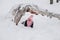 Distant photo of female kid looking into camera lying under tree log on snow and smiling wearing pink winter clothes in