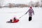 Distant photo of female Russian kid on sled with grandmother pulling granddaughter wearing pink winter clothes in park