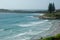 Distant surfers at a beach bordered by rock pools and pine trees.