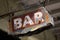 Distressed, rusty metal pole displays a faded sign that reads 'bar'.