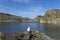 Disused quarry, blue sky with mounds of stone and cliffs with rocky outcrops. Person sitting at the water`s edge.