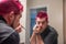 Diverse caucasian man with spiked pink hair applying eyeliner in the mirror