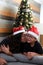 Divorced single mom and 7-year-old dark-skinned Latino son wear Santa hats in front of the Christmas tree to celebrate in solitude