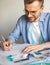 A DIY designer draws a sketch for a new costume project. A self-taught seamster with glasses is working at the table. A smiling