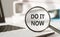 DO IT NOW - text on magnifying glass lying on laptop on office table