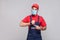 We do ontime. Young man with surgical medical mask in blue overall and red t-shirt standing and showing time on his wrist watch