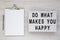 `Do what makes you happy` words on a lightbox, clipboard with blank sheet of paper on a white wooden background, top view.