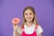 Do you want bite. Girl smiling face holds pink donut in hand, violet background. Kid smiling girl ready to bite donut