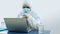 Doctor with blue mask and bioprotective suit working with his laptop in his office with his head resting on his hands with
