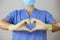 Doctor in a face mask and gloves forming a heart in front of blue shirt,