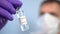 Doctor in a facial mask examines an ampoule with a coronavirus vaccine in his hand in protective gloves on a blue background