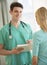 Doctor Holding Clipboard With Patient Standing In Foreground At